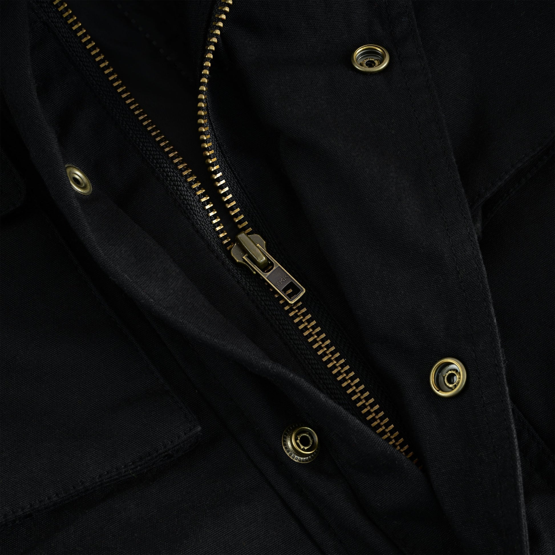 Extreme close-up shot of the heavy-duty brass main zipper and surrounding fabric on a black waxed cotton jacket, showing the quality of the hardware and a brass snap button closure.