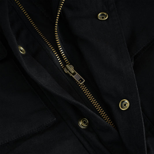 Extreme close-up shot of the heavy-duty brass main zipper and surrounding fabric on a black waxed cotton jacket, showing the quality of the hardware and a brass snap button closure.