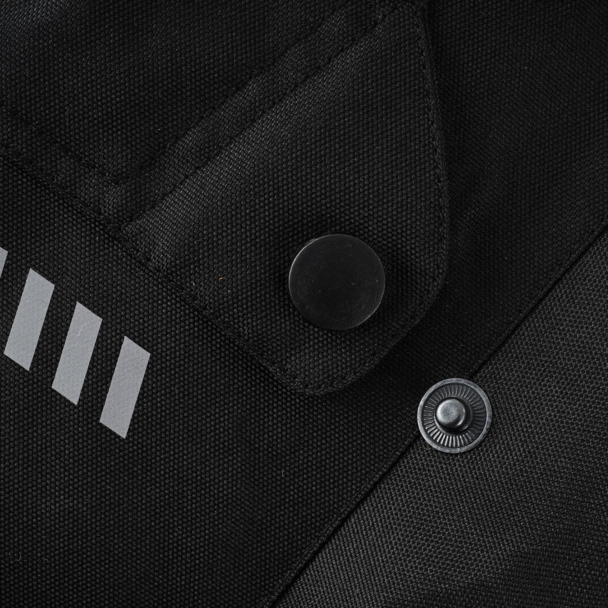 Extreme close-up of two snap buttons, one covered in black fabric, one metallic, positioned next to a series of silver reflective stripe accents on the jacket's black textile material.