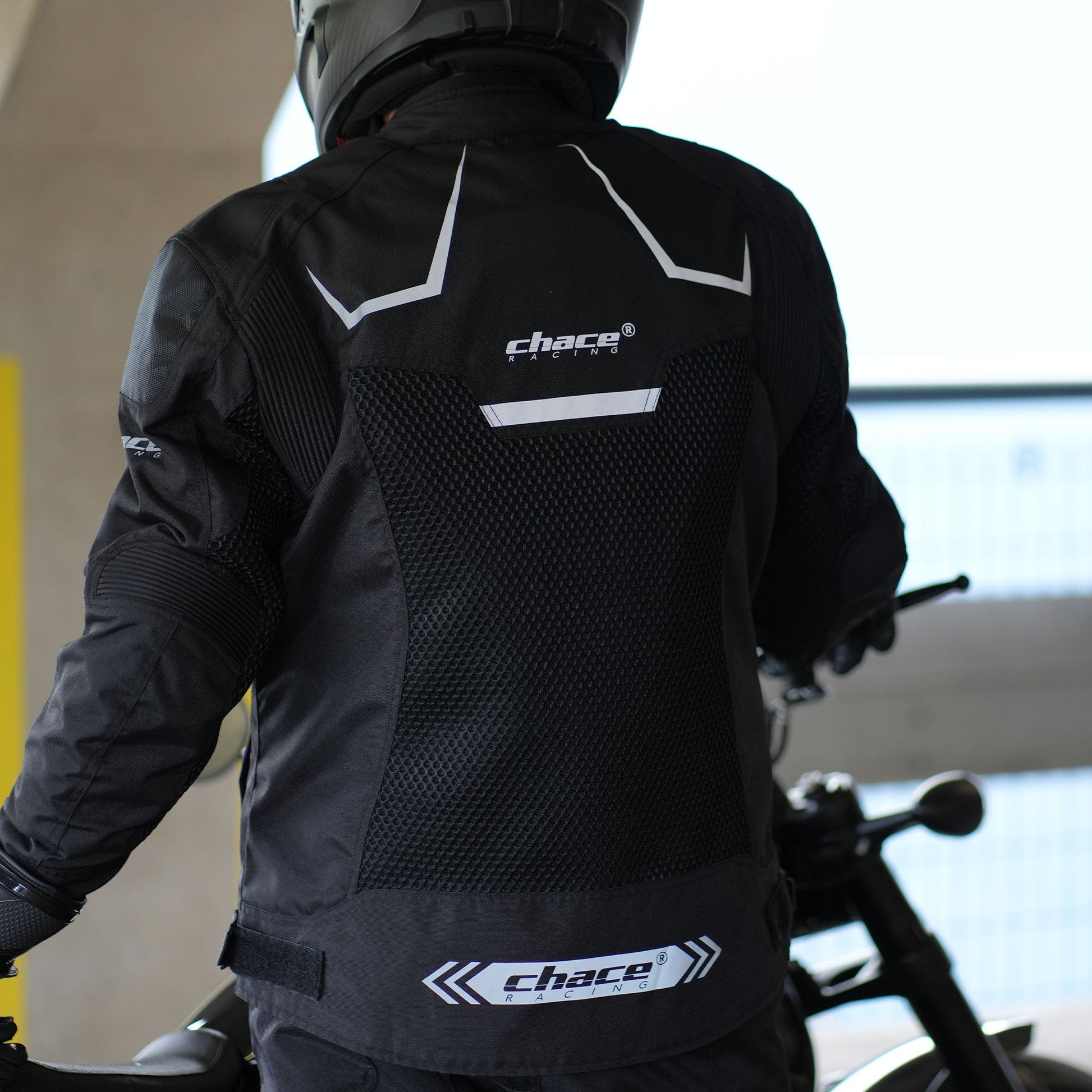 Rear view of a rider wearing the black Chace Volt motorcycle jacket, emphasizing the large breathable mesh back panel and the reflective "Chace Racing" logo strip at the lower hem.