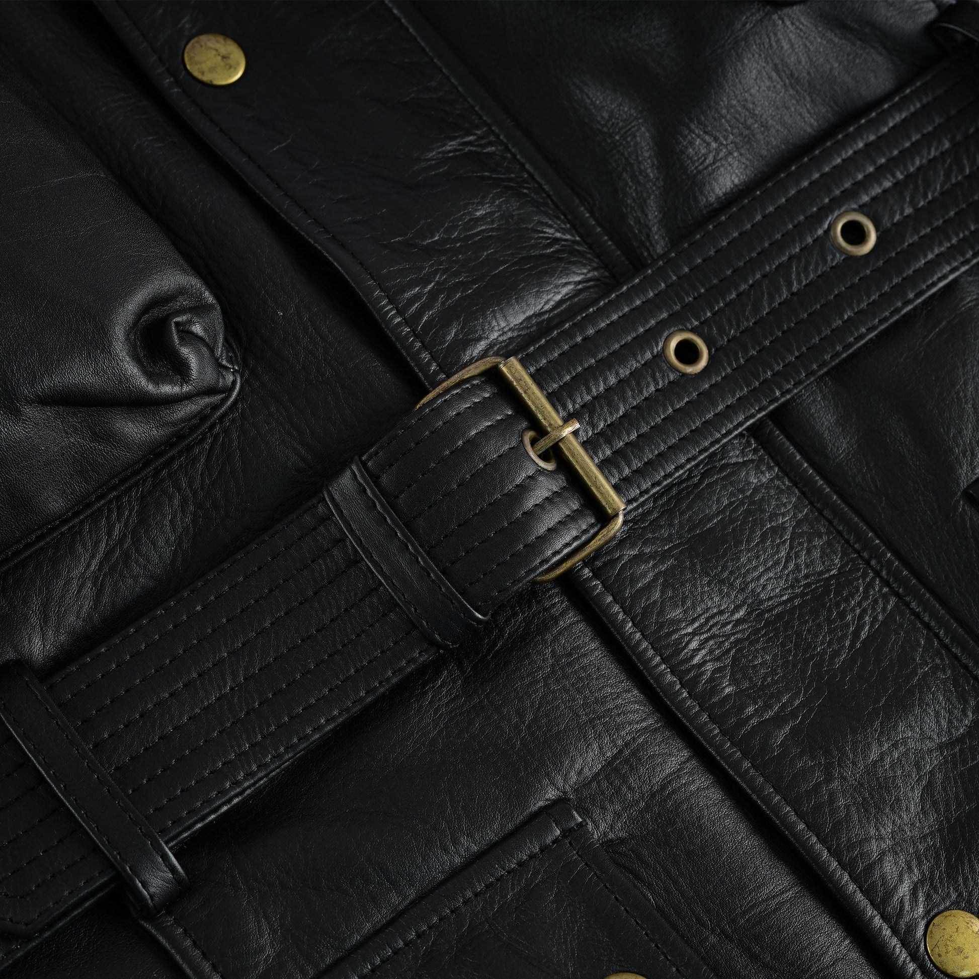 Extreme close-up of the jacket's leather waist belt, showing the stitched detailing, the adjustment holes, and the antique brass buckle and loop keeper.