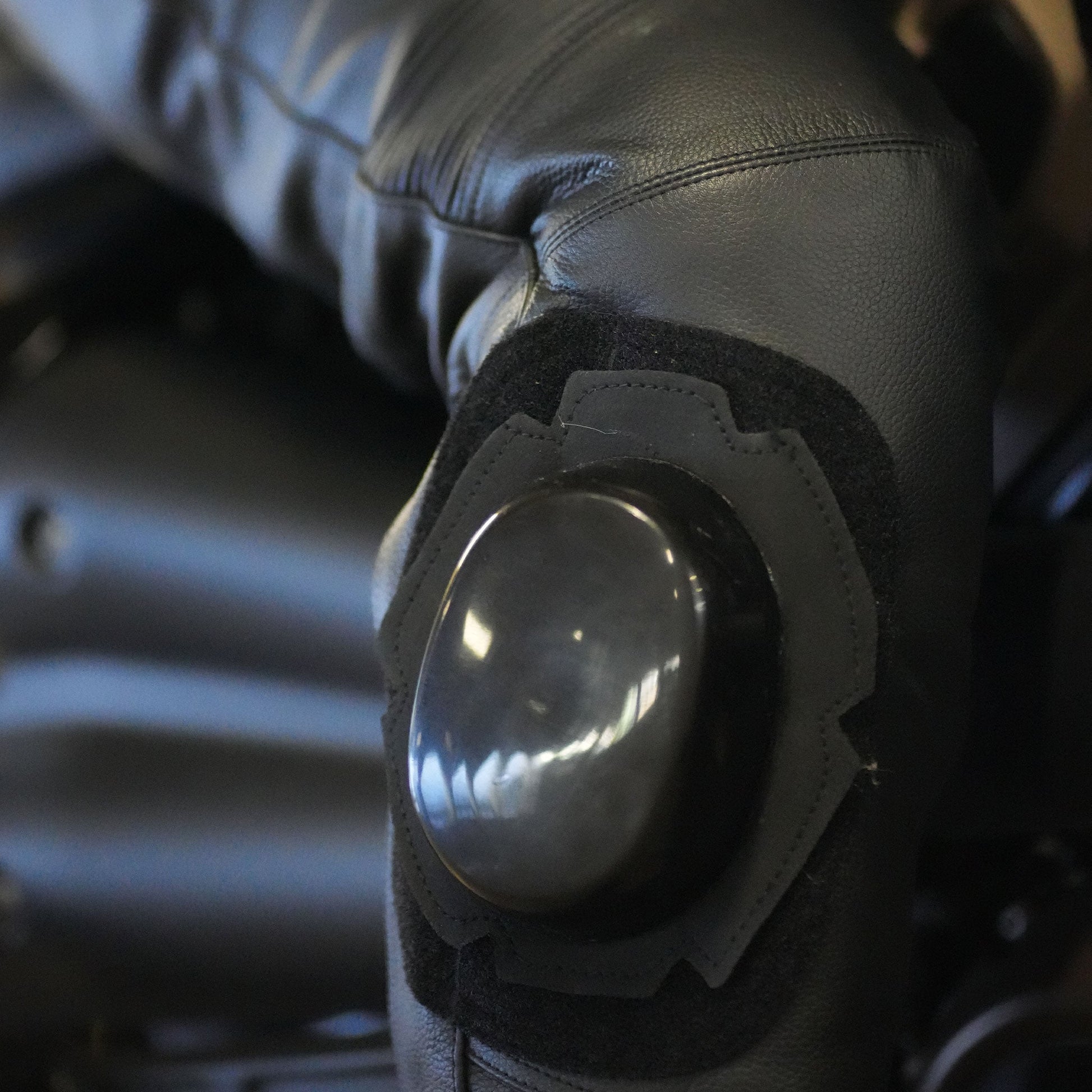 Extreme close-up of the glossy black, oval-shaped knee slider pad attached to the black leather motorcycle trousers via a hook-and-loop (Velcro) base.