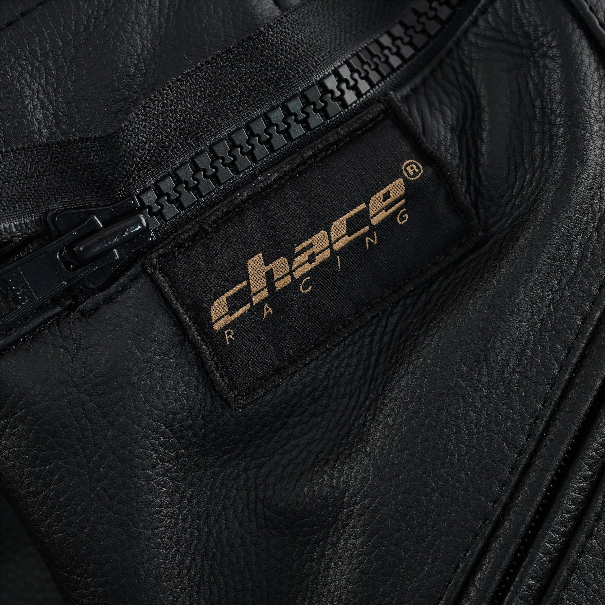 Close-up of the trousers' fly area, showing the black zipper and the black and gold stitched "Chace Racing" brand patch on the leather.