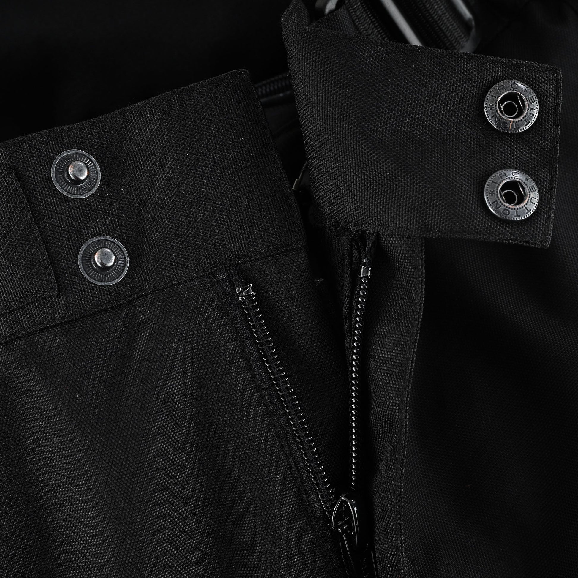 Close-up of the trousers' front fly closure, showing the partially open main zipper and the reinforced waist secured by dual metal snap buttons.