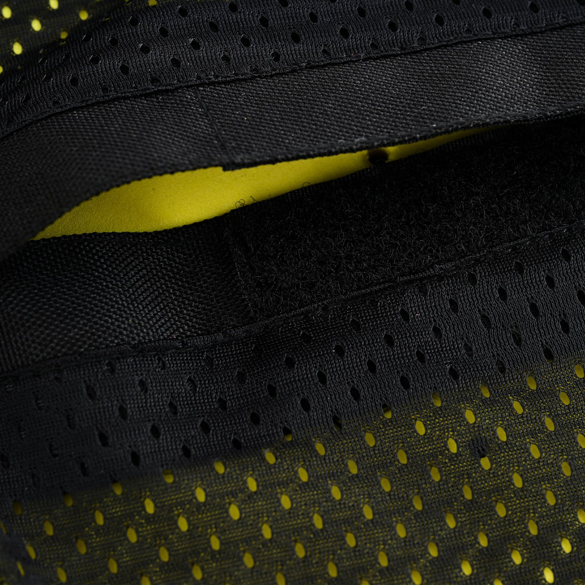 Extreme close-up of the opening flap for the back armor pocket, showing black hook-and-loop (Velcro) closures and a yellow interior through the perforated mesh lining, suggesting a high-visibility lining.