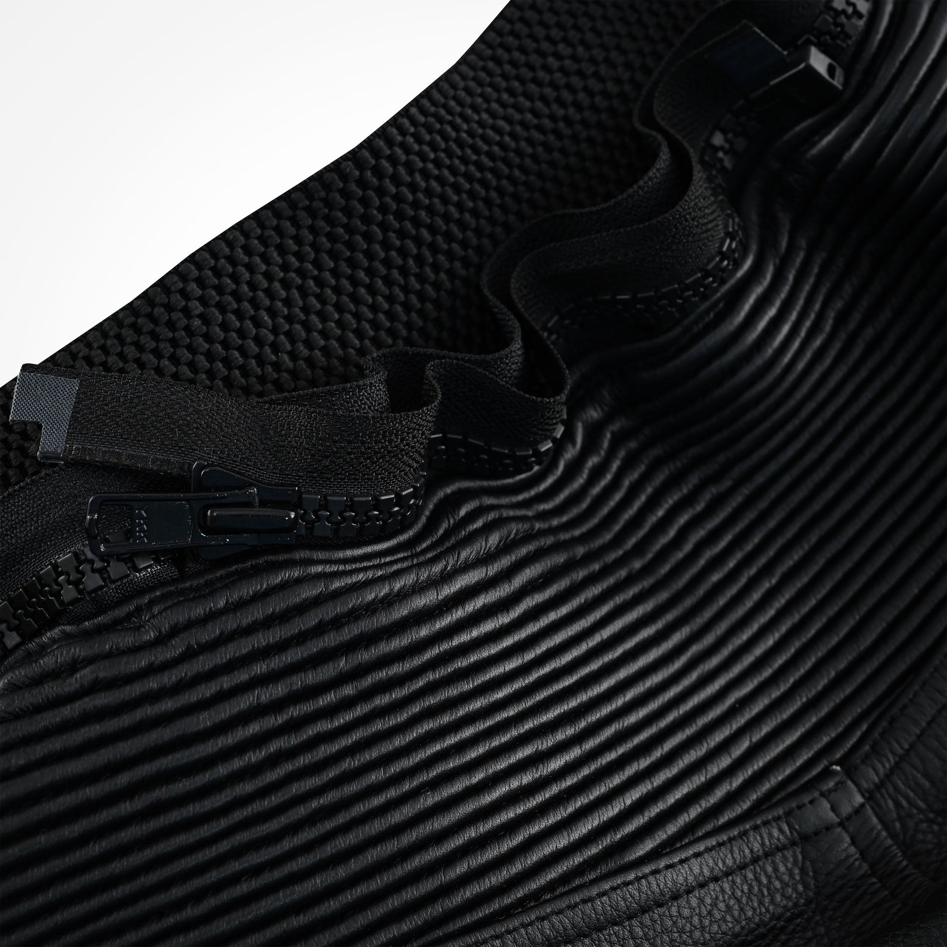 Close-up of the black leather trousers showing a heavy-duty zipper and pull tab interfacing with both the accordion stretch leather panel and a separate knit stretch fabric.
